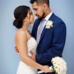 Bride and groom sharing a tender moment with bouquet against a blue background.