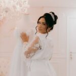 Bride in lace robe with wedding dress in elegant room, showcasing bridal elegance.